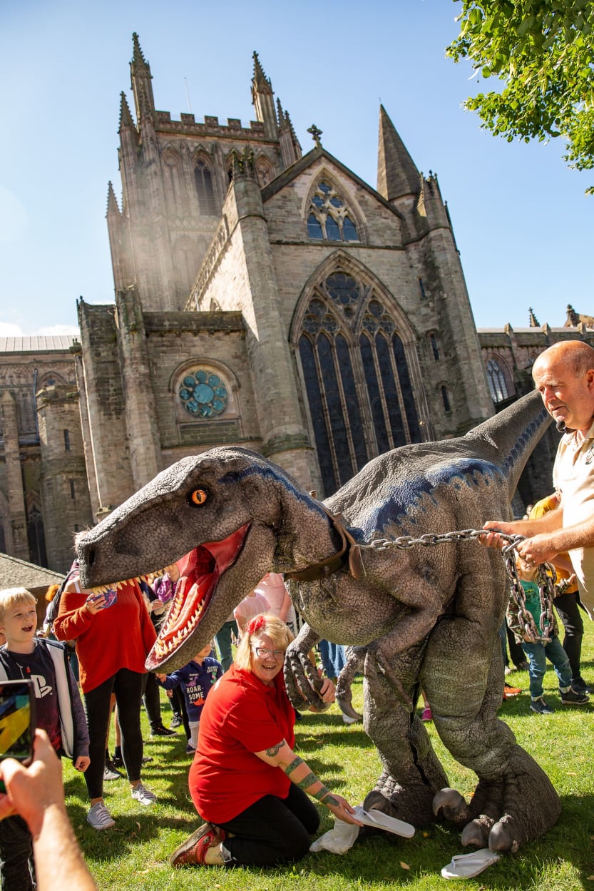 A lady in a red shirt is kneeling on the floor pretending to put a flip flip onto the foot of a dinosaur. The cathedral is in the background as well as a group of people in the sunshine