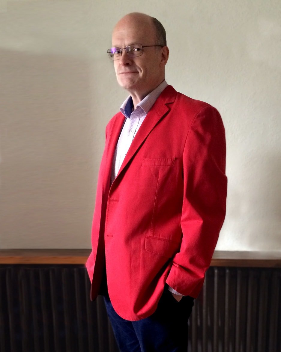 Tim stands smiling at the camera. He is wearing a red blazer and has his hand in one pocket