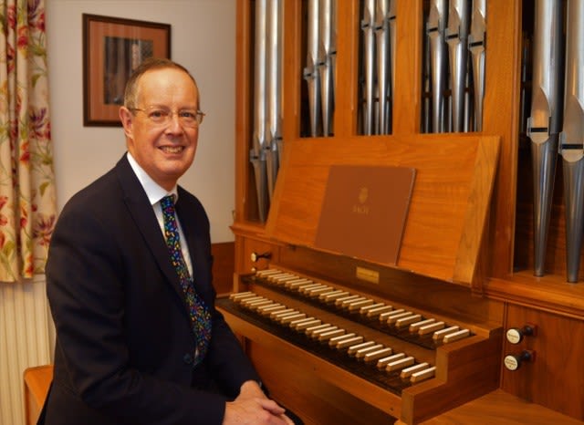 James Lancelot sitting in front of an organ. He is dressed in a suit and tie and smiling warmly at the camera