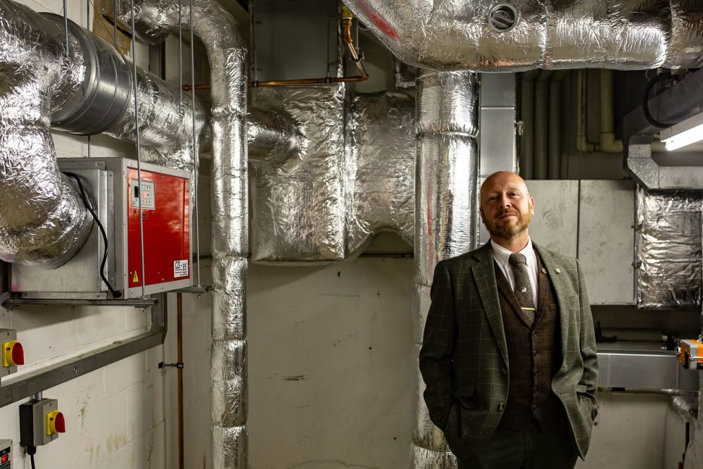 Alan is wearing a suit and tie and is smiling at the camera. He is standing in the plant room of the library and there are lots of pipes and technical equipment on the wall.