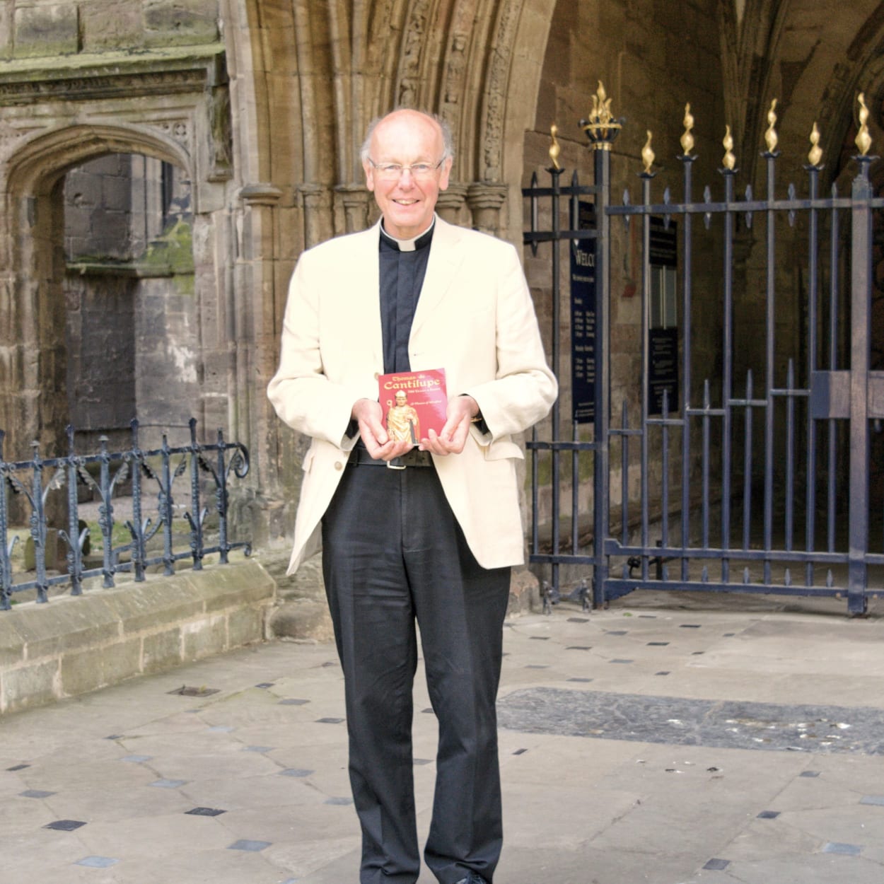The Dean stands outside the gates of the cathedral smiling with a copy of the book in his hands