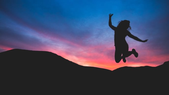 A silhouette of mountains at sunset with a person leaping for joy in front of them. Their silhouette is illuminated by a vibrant pink and purple sky