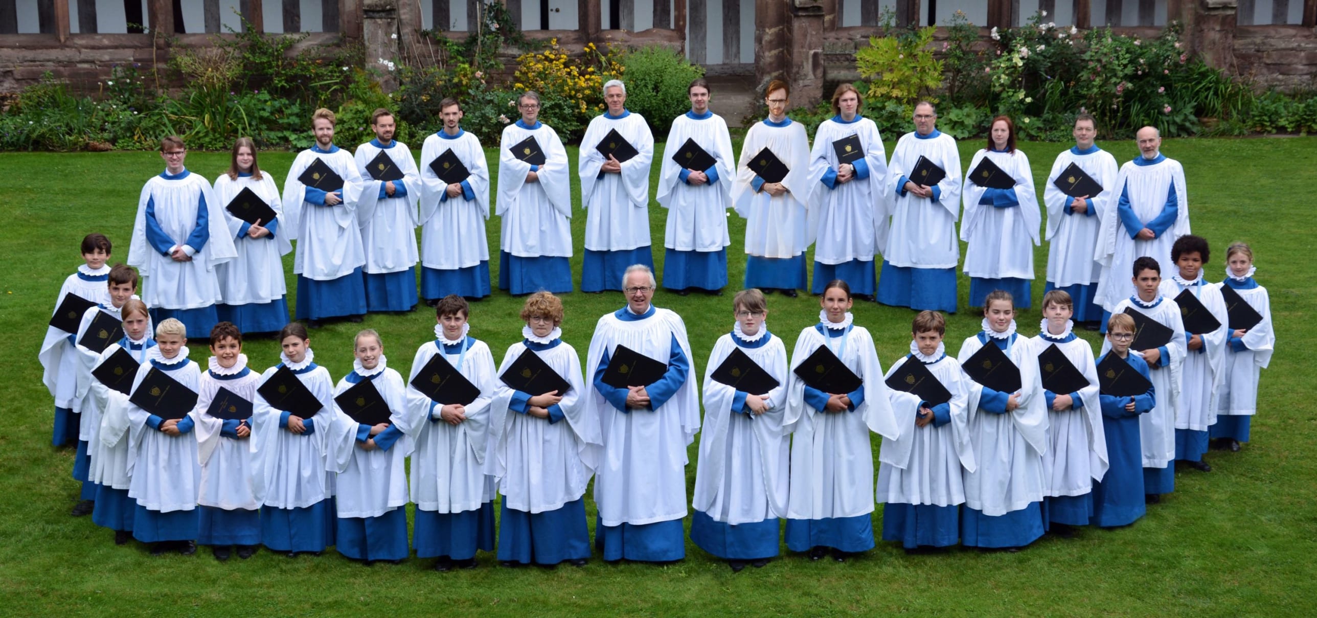 The choir of Hereford Cathedral stand in a circle in the Cloisters. They are wearing white and holding music scores in their arms. The choir consists of adults and children and they are all looking formally at the camera