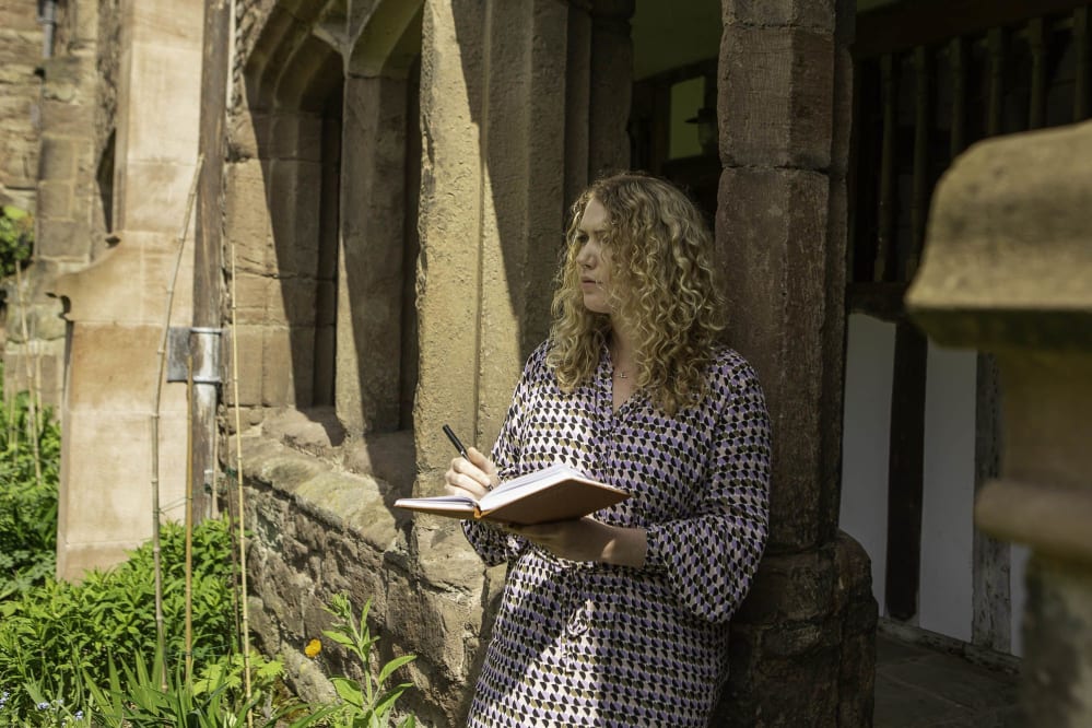 A woman stands in the Cloisters staring into the distance with a thoughtful look on her face. She is holding a pen and writing in an open sketchbook