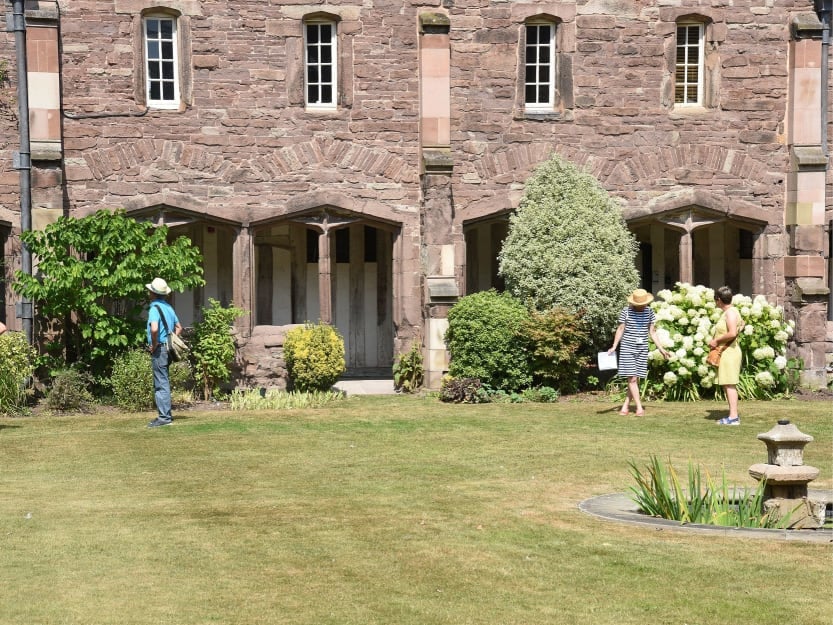 Visitors stand in the cloisters garden in bright sunshine. One is admiring shrubbery in the beds whilst another talks to a guide