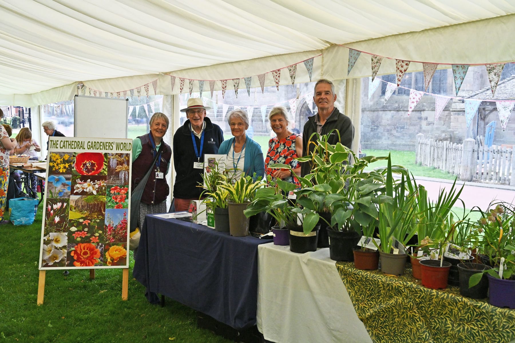 Five people are smiling at the camera inside the marquee. There is a table laden with plants in front of them